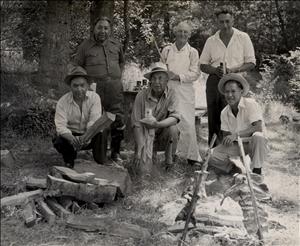 Six people stand and kneel in a grassy clearing in the forest. Most of them appear to be indigenous. They are kneeling in front of a small fire with fish cooking over the fire on sticks. One of them holds firewood. 